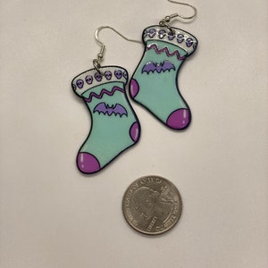 Puede incluir: Un par de pendientes colgantes con forma de calcet&iacute;n azul claro con un murci&eacute;lago morado y un borde de calaveras. Los pendientes son de metal y tienen un acabado plateado.