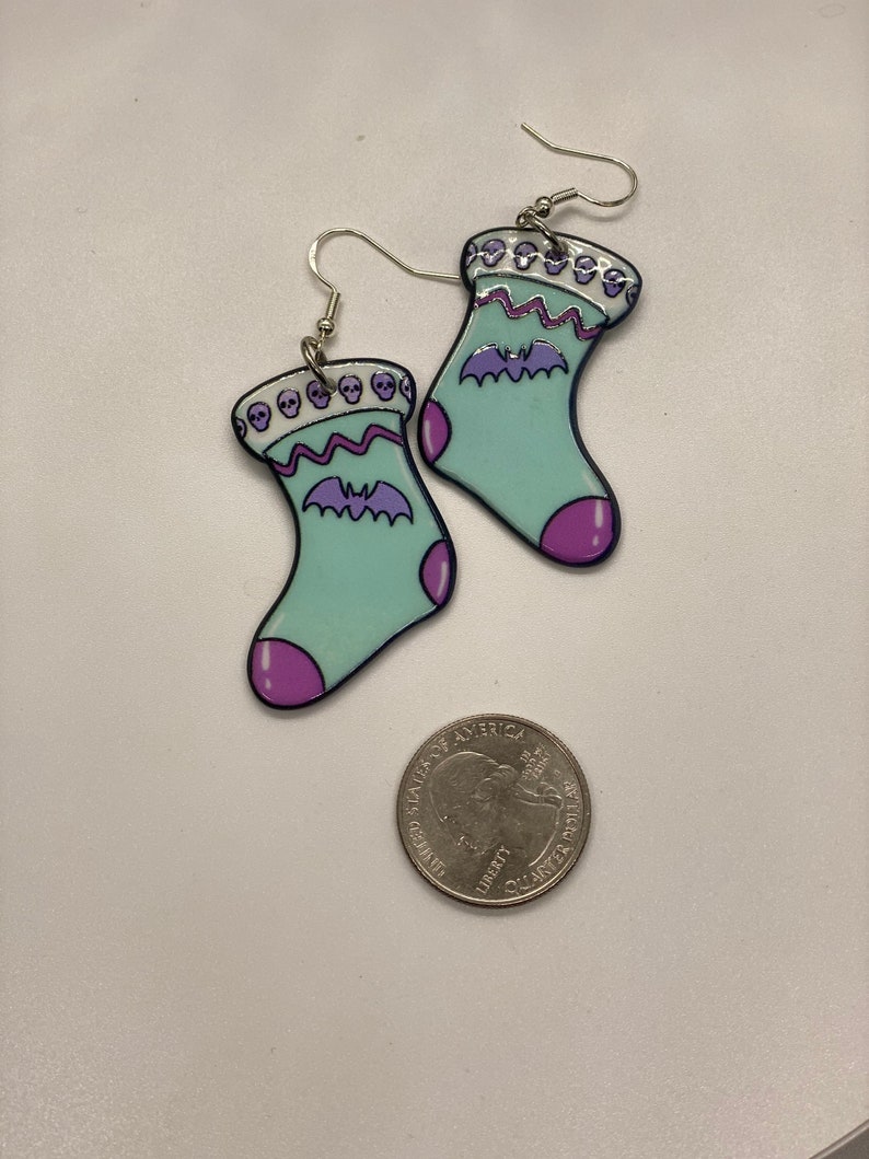 Puede incluir: Un par de pendientes colgantes con forma de calcet&iacute;n azul claro con un murci&eacute;lago morado y un borde de calaveras. Los pendientes son de metal y tienen un acabado plateado.