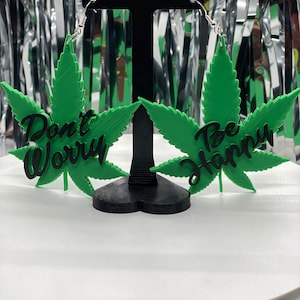 Peut inclure: Une paire de boucles d'oreilles en forme de feuille de marijuana verte avec des lettres noires. La boucle d'oreille gauche dit "Ne t'inquiète pas" et la boucle d'oreille droite dit "Sois heureux".