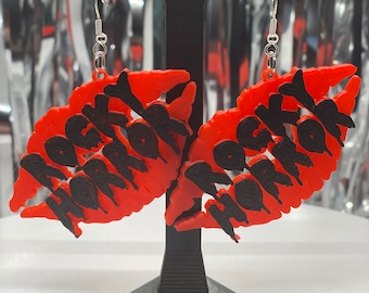 Puede incluir: Un par de pendientes rojos y negros. Los pendientes tienen forma de labios y presentan el texto "ROCKY HORROR" en negro. Los pendientes se exhiben sobre un soporte negro sobre un fondo plateado.