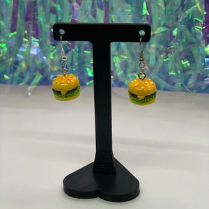 May include: A pair of dangle earrings featuring a miniature yellow and green hamburger charm. The earrings are displayed on a black heart-shaped stand.