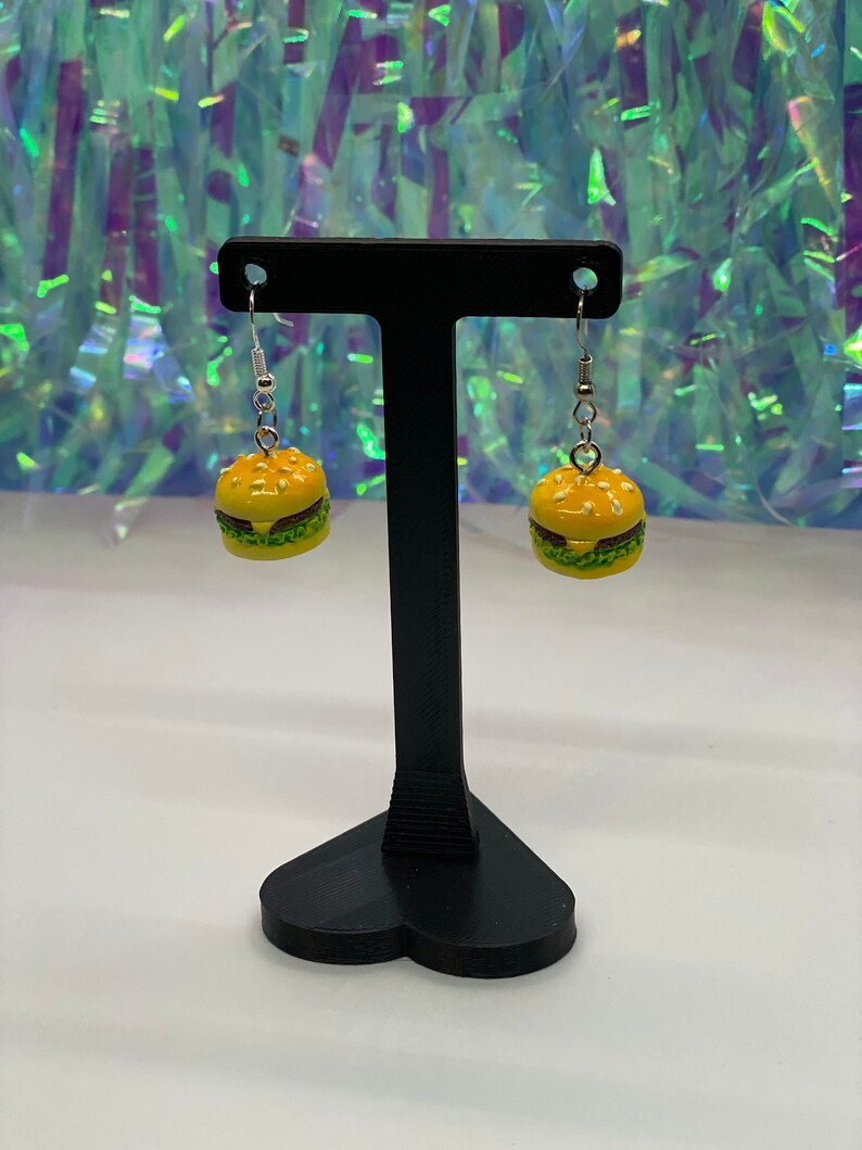 May include: A pair of dangle earrings featuring a miniature yellow and green hamburger charm. The earrings are displayed on a black heart-shaped stand.