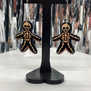 Puede incluir: Un par de pendientes de esqueleto con pintura negra y naranja. Los pendientes est&aacute;n colgados de un soporte negro en forma de coraz&oacute;n.