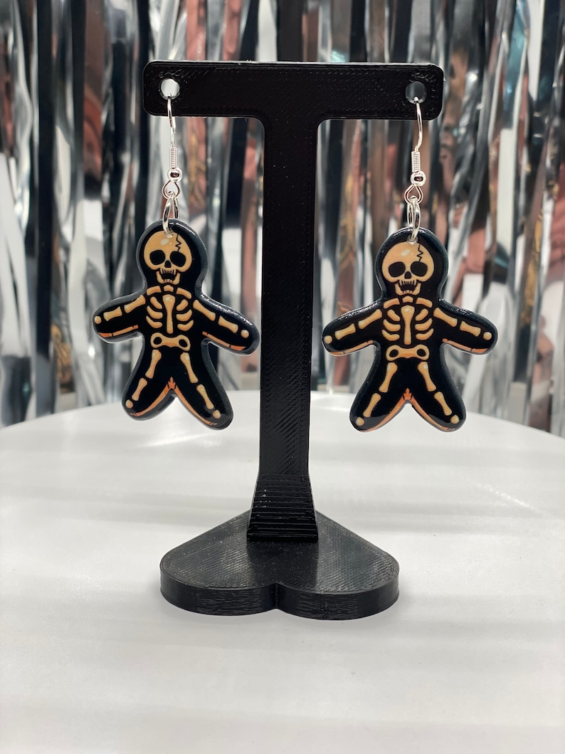 Puede incluir: Un par de pendientes de esqueleto con pintura negra y naranja. Los pendientes est&aacute;n colgados de un soporte negro en forma de coraz&oacute;n.