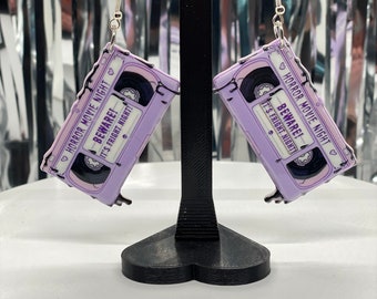 Puede incluir: Un par de pendientes de cassette púrpura con el texto "Horror Movie Night! Beware! It's Fright Night!" en cada pendiente.