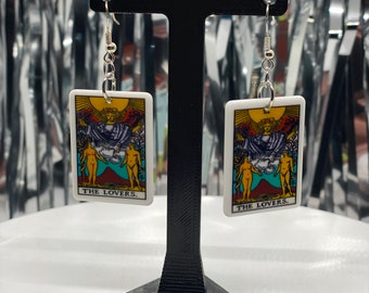 Puede incluir: Un par de pendientes de plata con dijes rectangulares blancos que representan la carta del tarot de Los Amantes. Los dijes tienen una ilustración colorida de dos figuras de pie con una figura alada sobre ellas. El texto "THE LOVERS." está impreso debajo de la ilustración.