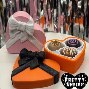 May include: Three heart-shaped boxes: pink with a white bow, orange with a black bow, and orange with chocolates inside. The chocolates have various toppings. The boxes are on a white surface with a silver and pink backdrop. The text "PRETTY UNDEAD" is on the bottom right.