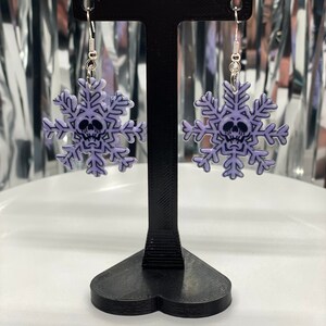 Puede incluir: Un par de pendientes de copo de nieve plateados con un dise&ntilde;o de calavera morada. Los pendientes est&aacute;n colgados de un soporte para pendientes negro.