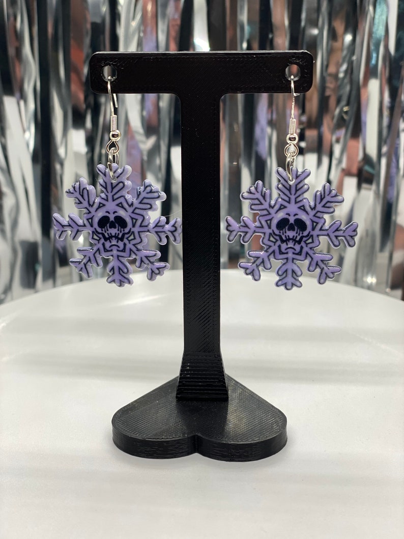 Puede incluir: Un par de pendientes de copo de nieve plateados con un dise&ntilde;o de calavera morada. Los pendientes est&aacute;n colgados de un soporte para pendientes negro.