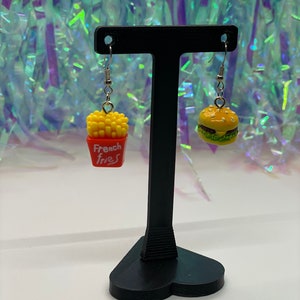 May include: A pair of dangle earrings with a burger and french fries charm. The burger charm is yellow with a green lettuce and red tomato. The french fries charm is yellow with a red box that says "French Fries". The earrings are hanging from a black heart-shaped stand.