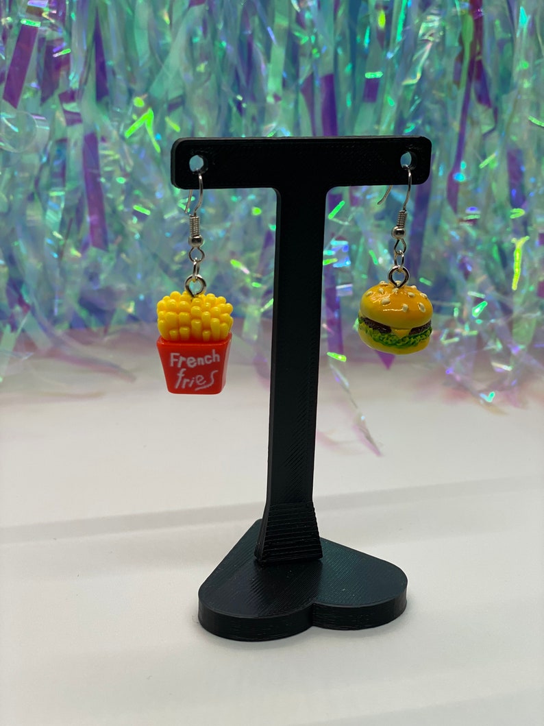 May include: A pair of dangle earrings with a burger and french fries charm. The burger charm is yellow with a green lettuce and red tomato. The french fries charm is yellow with a red box that says "French Fries". The earrings are hanging from a black heart-shaped stand.