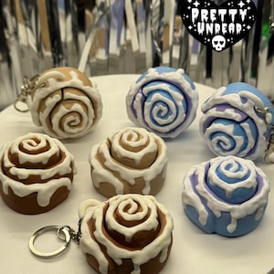 Puede incluir: Una colección de llaveros con forma de rollo de canela en varios colores, incluyendo marrón, beige y azul, con detalles de glaseado blanco. Algunos llaveros tienen anillos plateados. La imagen también presenta un logotipo en forma de corazón con el texto "PRETTY UNDEAD".