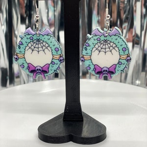Puede incluir: Un par de pendientes de plata con un dise&ntilde;o de corona verde, morado y blanco. La corona tiene una telara&ntilde;a, una calavera y un lazo.