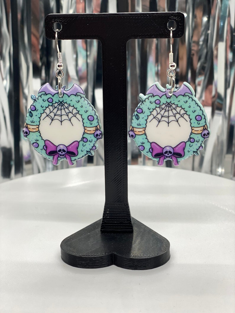 Puede incluir: Un par de pendientes de plata con un dise&ntilde;o de corona verde, morado y blanco. La corona tiene una telara&ntilde;a, una calavera y un lazo.