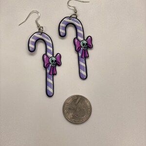 Puede incluir: Un par de pendientes de caramelo de ca&ntilde;a rayados en morado y blanco con un lazo morado y una calavera blanca en cada uno. Los pendientes son plateados y tienen un cierre de gancho.