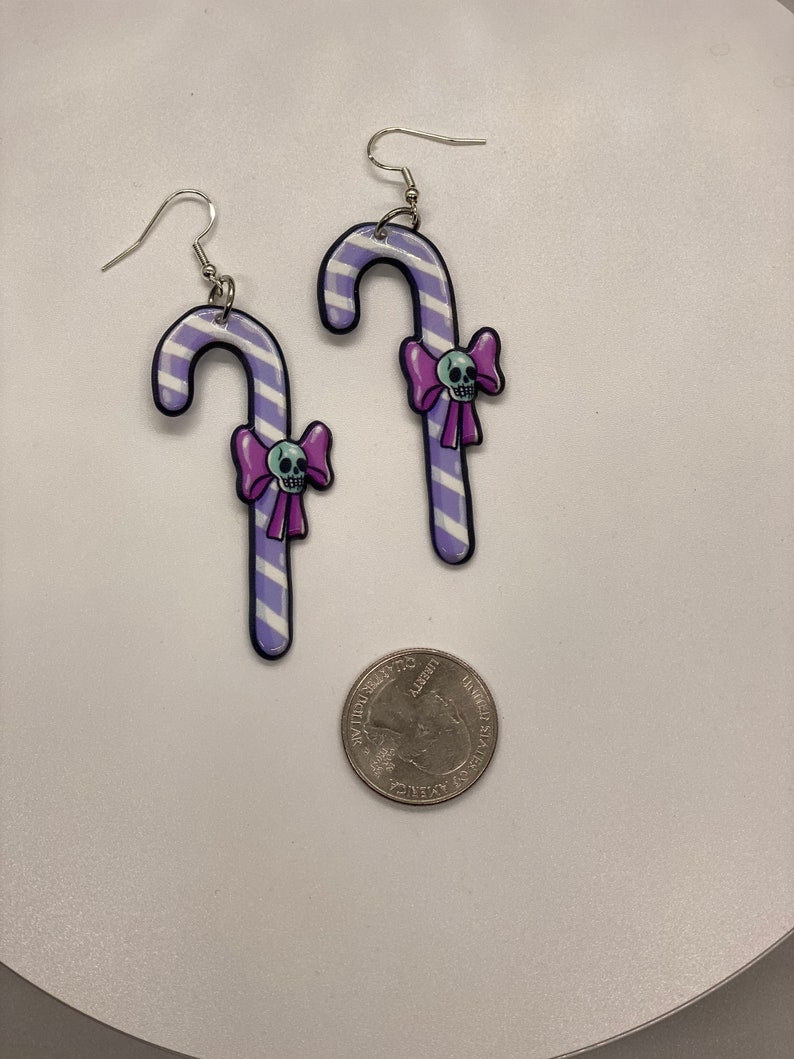 Puede incluir: Un par de pendientes de caramelo de ca&ntilde;a rayados en morado y blanco con un lazo morado y una calavera blanca en cada uno. Los pendientes son plateados y tienen un cierre de gancho.