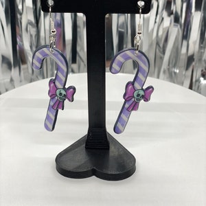 Puede incluir: Un par de pendientes de caramelo de ca&ntilde;a rayados en morado y blanco con un lazo rosa y un dije de calavera colgando de cada uno. Los pendientes se muestran en un soporte negro en forma de coraz&oacute;n.