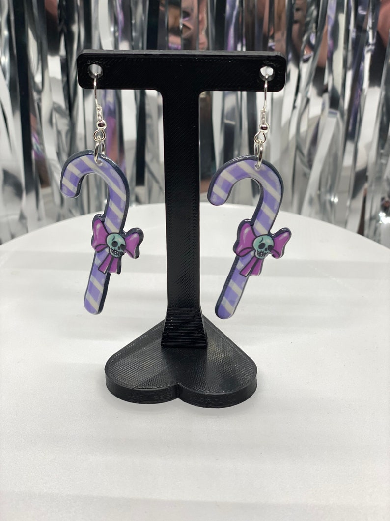 Puede incluir: Un par de pendientes de caramelo de ca&ntilde;a rayados en morado y blanco con un lazo rosa y un dije de calavera colgando de cada uno. Los pendientes se muestran en un soporte negro en forma de coraz&oacute;n.