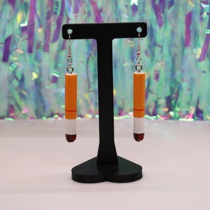 Op de afbeelding: Een paar hangende oorbellen in de vorm van sigaretten. De oorbellen zijn oranje, wit en rood. Ze hangen aan een zwarte oorbellenstandaard.