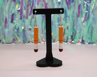 Puede incluir: Un par de pendientes colgantes con forma de cigarrillos. Los pendientes son de color naranja, blanco y rojo. Están colgados de un soporte de pendientes negro.