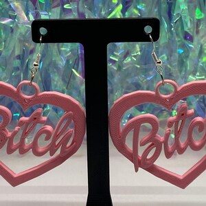 May include: A pair of pink heart-shaped earrings with the word "Bitch" written in a cursive font inside each heart.