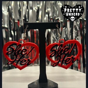May include: A pair of red heart-shaped earrings with the text "She/Her" in black lettering.