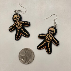 Puede incluir: Un par de pendientes colgantes con un dise&ntilde;o de esqueleto en blanco y negro. El esqueleto lleva un atuendo negro y tiene una calavera blanca. Los pendientes est&aacute;n hechos de metal y tienen un gancho plateado.