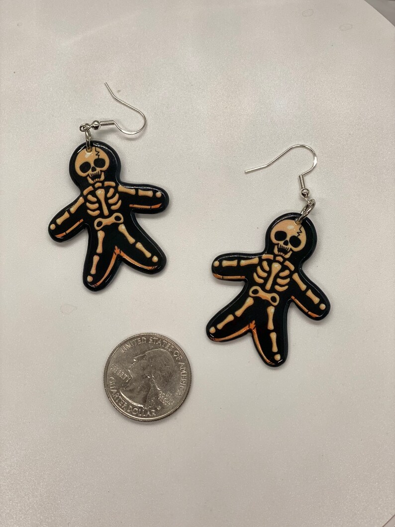 Puede incluir: Un par de pendientes colgantes con un dise&ntilde;o de esqueleto en blanco y negro. El esqueleto lleva un atuendo negro y tiene una calavera blanca. Los pendientes est&aacute;n hechos de metal y tienen un gancho plateado.