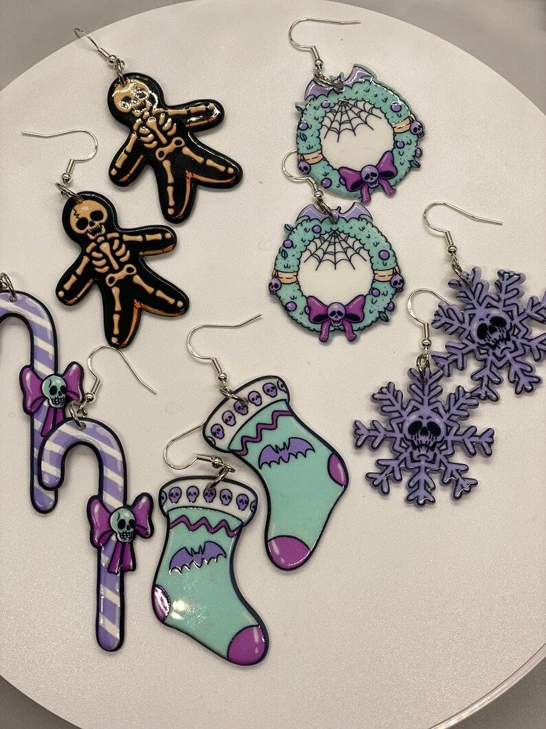 Puede incluir: Un conjunto de 10 pendientes de Navidad espeluznantes con una variedad de dise&ntilde;os, que incluyen una ca&ntilde;a de az&uacute;car, una media, un copo de nieve, una corona y un esqueleto. Los pendientes est&aacute;n hechos de pl&aacute;stico y tienen una combinaci&oacute;n de colores p&uacute;rpura, verde y negro.