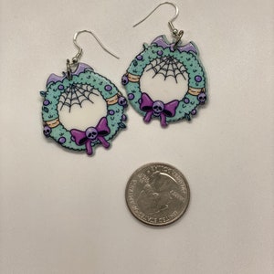 Puede incluir: Un par de pendientes de plata con un dise&ntilde;o de corona verde. La corona est&aacute; decorada con una telara&ntilde;a blanca, un lazo morado con una calavera y peque&ntilde;as calaveras.