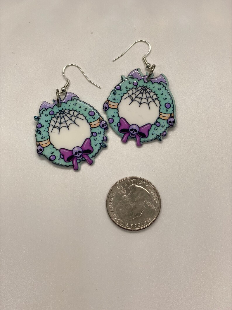 Puede incluir: Un par de pendientes de plata con un dise&ntilde;o de corona verde. La corona est&aacute; decorada con una telara&ntilde;a blanca, un lazo morado con una calavera y peque&ntilde;as calaveras.