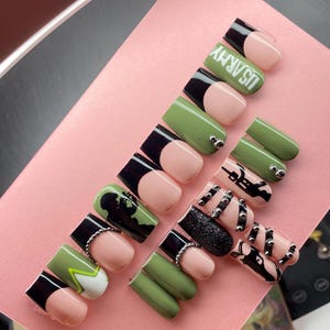 Op de afbeelding: Een verzameling persnagels in verschillende ontwerpen. Sommige nagels zijn olijfgroen, zwart en nude, met zilveren studs en de tekst "US ARMY". Andere nagels hebben zwart-witte patronen en glitter.
