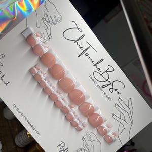 May include: A set of 10 press-on nails with a white and pink design. The nails are arranged in two rows on a white card with the text "Chic Touche By Sav" and "Luxury press on nails".
