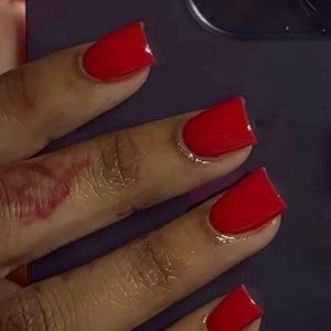 Könnte beinhalten: Nahaufnahme einer Hand mit leuchtend rotem Nagellack auf den Fingernägeln. Die Nägel sind sauber geformt und poliert und reflektieren das Licht. Der Hintergrund ist eine verschwommene lila Oberfläche.