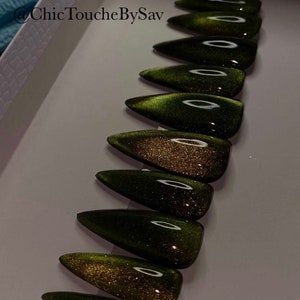 May include: A collection of almond-shaped, press-on nails in a dark green colour with a shimmering, cat-eye effect. The nails are arranged in a row, displaying their glossy finish. The text "@Chic ToucheBySav" is visible in the upper left corner.