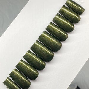 May include: A set of ten olive green press-on nails. Each nail has a glossy finish with a subtle shimmer and a white stripe near the cuticle. The nails are arranged diagonally on a white surface.