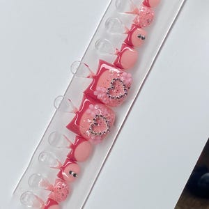 Puede incluir: Un conjunto de uñas postizas para los pies rosas y brillantes. Las uñas presentan una variedad de diseños, incluyendo rosa liso, purpurina y adornos en forma de corazón con pedrería. Las uñas están dispuestas en una pantalla de plástico transparente.