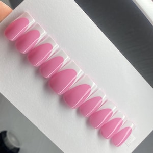 Pode incluir: Um conjunto de unhas postiças rosa com uma borda transparente e arredondada. As unhas estão dispostas em uma fileira sobre uma superfície branca, exibindo uma cor rosa sólida e brilhante. As unhas têm formato oval.