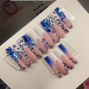 Puede incluir: Un conjunto de uñas artificiales con varios diseños. Algunas uñas presentan una base blanca con estampado de leopardo azul, otras una base rosa con manchas de leopardo azules, y algunas un efecto degradado azul. Se añaden pedrería azul para mayor brillo.