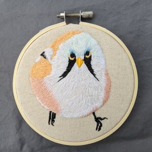 Könnte beinhalten: Stickerei in einem runden Rahmen mit einem weißen, rosa und blauen Vogel mit schwarzen Augen und einem gelben Schnabel. Der Vogel sitzt auf einem beigefarbenen Stoffhintergrund.