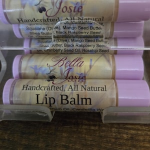 May include: Stacked lip balm tubes with light purple caps and labels. The labels feature the text "Bella Josie" and "Handcrafted All Natural Lip Balm". Ingredients include plum oil, shea butter, and black raspberry seed.