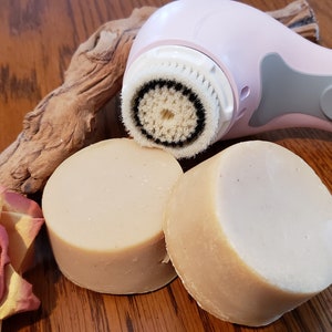 Puede incluir: Dos barras redondas de jabón natural, una beige y otra marrón claro, junto a un cepillo eléctrico de limpieza facial rosa y gris con un cabezal de cepillo de cerdas blancas.