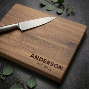 May include: A rectangular wooden cutting board with a stainless steel knife. The board is engraved with the name "ANDERSON" and the year "EST. 2026". The wood has a natural grain and a warm brown color.