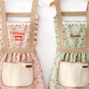 May include: Two floral aprons, one pink and one green, are displayed. The aprons feature beige pockets, straps, and trim. The pink apron is embroidered with the name "Lillian", while the green apron has "Brianna" embroidered on it.