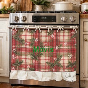 May include: A red, green and cream plaid oven towel hanging on a stainless steel stove handle. The towel features a repeating pattern of pine branches and white snowflakes, with the name Mavis embroidered in green and a white ruffled hem.