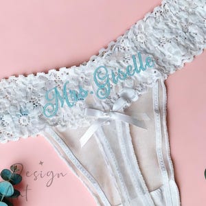 Puede incluir: Braga tanga de encaje blanco con el texto "Mrs. Giselle" en escritura azul claro. La ropa interior tiene un pequeño lazo y está colocada sobre un fondo rosa con ramitas de hojas azul-verdes.
