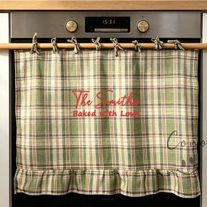 May include: A green and white plaid fabric oven curtain with a ruffled bottom edge, hanging from a wooden rod. The fabric features red embroidered text that reads The Smiths Baked with Love. The curtain is positioned over a stainless steel oven.