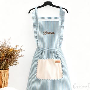 May include: A blue and white striped apron with the name "Brianna" embroidered on the chest. The apron has a large beige pocket and ruffled edges. It hangs on a black hanger against a white wall.