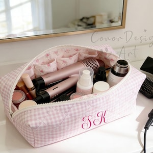 Puede incluir: Un neceser rosa y blanco a cuadros con las iniciales "SK" bordadas. La bolsa está abierta, revelando varios productos de belleza, incluyendo barras de labios, secadores de pelo y productos para el cuidado de la piel. El interior presenta bolsillos con estampado floral.