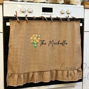 May include: A brown and white gingham oven towel with a ruffled bottom edge, featuring an embroidered bouquet of yellow sunflowers and the text The Michelle in black script, attached to the oven handle with brown gingham fabric ties.